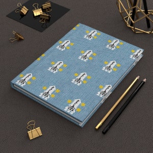 May include: A blue notebook with a repeating pattern of a white rocket ship and yellow stars. The notebook is lying on a dark gray surface with two pencils and a gold paperclip.