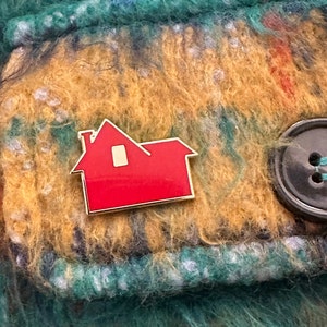May include: A red house-shaped enamel pin with a gold border and a small white window. The pin is set against a textured background of teal, gold, and brown fabric, with a large black button.