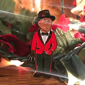 May include: Enamel pin featuring a character with a scarred face, wearing a red tuxedo jacket, green pants, and a black hat. The pin has a brown glove with sharp claws. The pin is set against a backdrop of green and red decorative elements.
