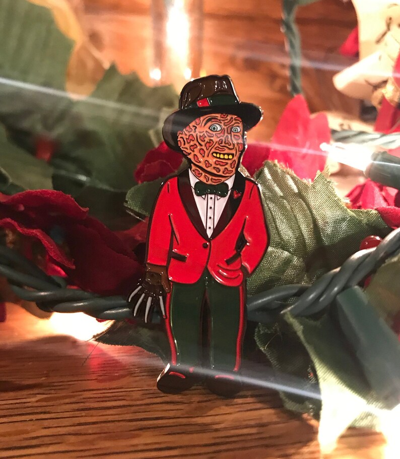 May include: Enamel pin featuring a character with a scarred face, wearing a red tuxedo jacket, green pants, and a black hat. The pin has a brown glove with sharp claws. The pin is set against a backdrop of green and red decorative elements.
