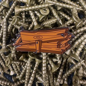 May include: A brown and orange enamel pin depicting a stack of wooden planks with nails. The pin is set against a background of gold-colored screws.
