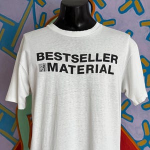 May include: A white short-sleeved t-shirt featuring the black text "BESTSELLER MATERIAL" printed across the chest. The design includes a small graphic icon of a person walking inside a square box positioned to the left of the word "MATERIAL."