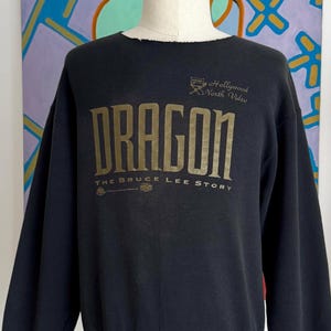 May include: Black sweatshirt featuring gold lettering that reads "DRAGON THE BRUCE LEE STORY". The sweatshirt has long sleeves and a crew neck. The text "By Hollywood North Wales" is above the word "DRAGON".