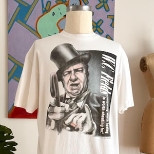May include: White t-shirt featuring a black and white graphic of W.C. Fields in a top hat, holding a cigar and pointing. The text "W.C. Fields" and "is worth cheating for" are printed on the right side of the shirt. The shirt is displayed on a mannequin.