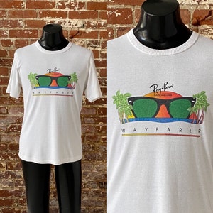 May include: White t-shirt with a graphic of Ray-Ban Wayfarer sunglasses with a sunset and palm trees. The text "Ray-Ban" and "Wayfarer" are printed on the shirt.