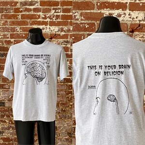 May include: Grey t-shirt with a black print on the front that says "This is your brain on science" with a diagram of a brain and scientific formulas. The back of the shirt says "This is your brain on religion" with a simple drawing of a brain and the word "DUHH!!"