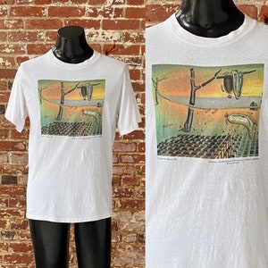 May include: White t-shirt with a graphic print of Salvador Dali's "The Persistence of Memory". The painting features a melting clock, a tree, and a desert landscape.