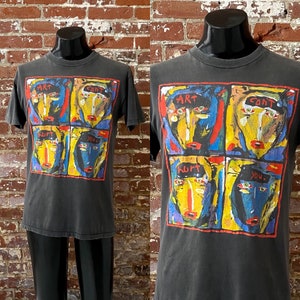 May include: Dark gray t-shirt with a colorful graphic of four stylized faces. Each face is framed in a red border and features bold colors like yellow, blue, and red. The words "ART", "HURT", and "YOU" are also visible.