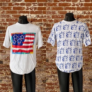 May include: Two white t-shirts on mannequins. One shirt features a colorful American flag design with stars and stripes. The other shirt has a repeating blue and white pattern of the Statue of Liberty.