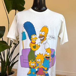 May include: White t-shirt featuring a colorful graphic of the Simpson family. The design includes Marge, Homer, Bart, Lisa, and Maggie Simpson in a living room setting. Homer says "Say Chee".
