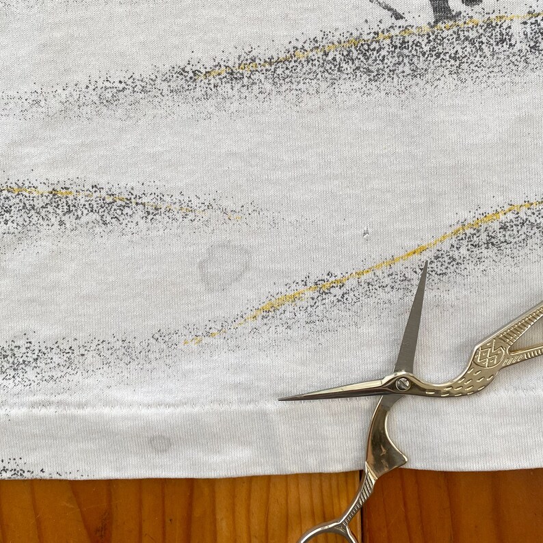 Puede incluir: Camiseta blanca con un dise&ntilde;o abstracto descolorido con detalles en gris y amarillo. La camiseta tiene cuello redondo y mangas cortas. Unas tijeras doradas est&aacute;n en primer plano, con las hojas apuntando hacia la camiseta.