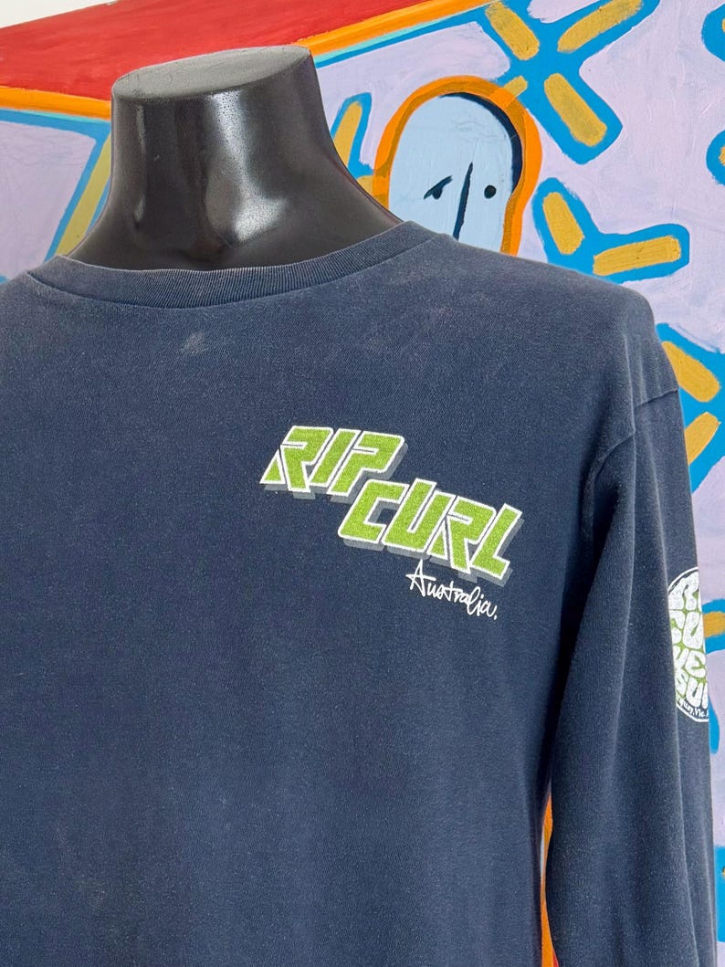 May include: Navy blue long-sleeve shirt with the Rip Curl logo in green and white on the chest. The word "Australia" is printed below the logo. A smaller logo is visible on the sleeve.