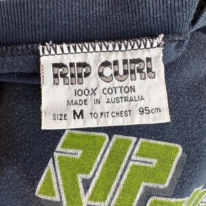 May include: Close-up of a navy blue t-shirt with a white tag. The tag reads "RIP CURL 100% COTTON MADE IN AUSTRALIA SIZE M TO FIT CHEST 95cm". The shirt has a green and white graphic design.