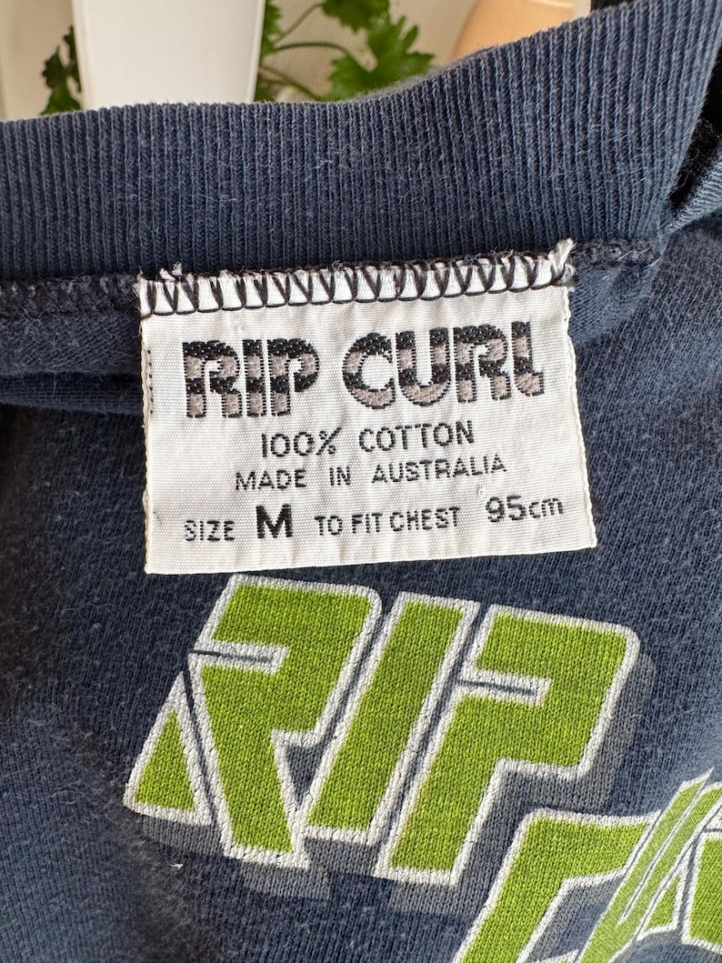 May include: Close-up of a navy blue t-shirt with a white tag. The tag reads "RIP CURL 100% COTTON MADE IN AUSTRALIA SIZE M TO FIT CHEST 95cm". The shirt has a green and white graphic design.