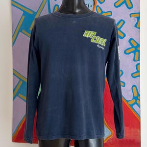 May include: A navy blue long-sleeve shirt with the text "RIP CURL" in green and white on the chest. The shirt has a crew neck and the word "Australia" is printed below the logo. The shirt is displayed on a black mannequin.