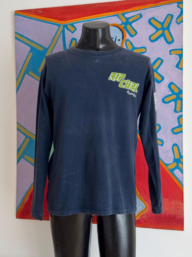 May include: A navy blue long-sleeve shirt with the text "RIP CURL" in green and white on the chest. The shirt has a crew neck and the word "Australia" is printed below the logo. The shirt is displayed on a black mannequin.