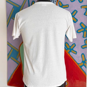 Może przedstawiać: Gładki, biały t-shirt z kr&oacute;tkim rękawem, prezentowany na czarnym manekinie. Koszulka wykonana jest z lekkiego materiału. Tło przedstawia kolorowy abstrakcyjny obraz z czerwonymi, fioletowymi i niebieskimi odcieniami.