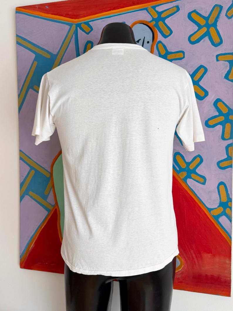 Może przedstawiać: Gładki, biały t-shirt z kr&oacute;tkim rękawem, prezentowany na czarnym manekinie. Koszulka wykonana jest z lekkiego materiału. Tło przedstawia kolorowy abstrakcyjny obraz z czerwonymi, fioletowymi i niebieskimi odcieniami.