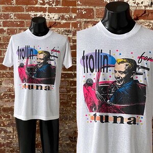 May include: White t-shirt with a colorful graphic of a person driving a car. The graphic says "trollin' for tuna".