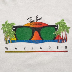 May include: White t-shirt with a graphic of Ray-Ban Wayfarer sunglasses in front of a sunset with palm trees. The text "Ray-Ban" is above the sunglasses and "BAUSCH & LOMB" is below. The text "WAYFARER" is below the sunglasses.