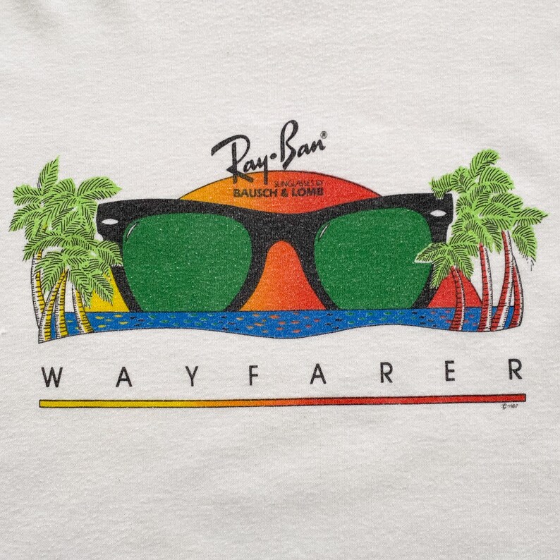 May include: White t-shirt with a graphic of Ray-Ban Wayfarer sunglasses in front of a sunset with palm trees. The text "Ray-Ban" is above the sunglasses and "BAUSCH & LOMB" is below. The text "WAYFARER" is below the sunglasses.