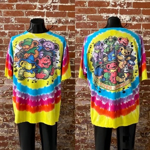 Op de afbeelding: Tie-dye T-shirt met een kleurrijke illustratie van cartoonberen die muziekinstrumenten bespelen. Het shirt heeft een regenboog tie-dye patroon met gele, blauwe, roze en oranje kleuren. De tekst "Grateful Dead" is op het shirt gedrukt.