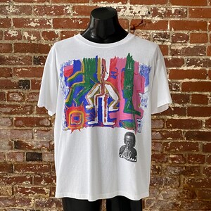 May include: White t-shirt with a colorful abstract design featuring a sun, figures, and lines. A black and white portrait of a man with the word "miles" written below is in the bottom right corner.