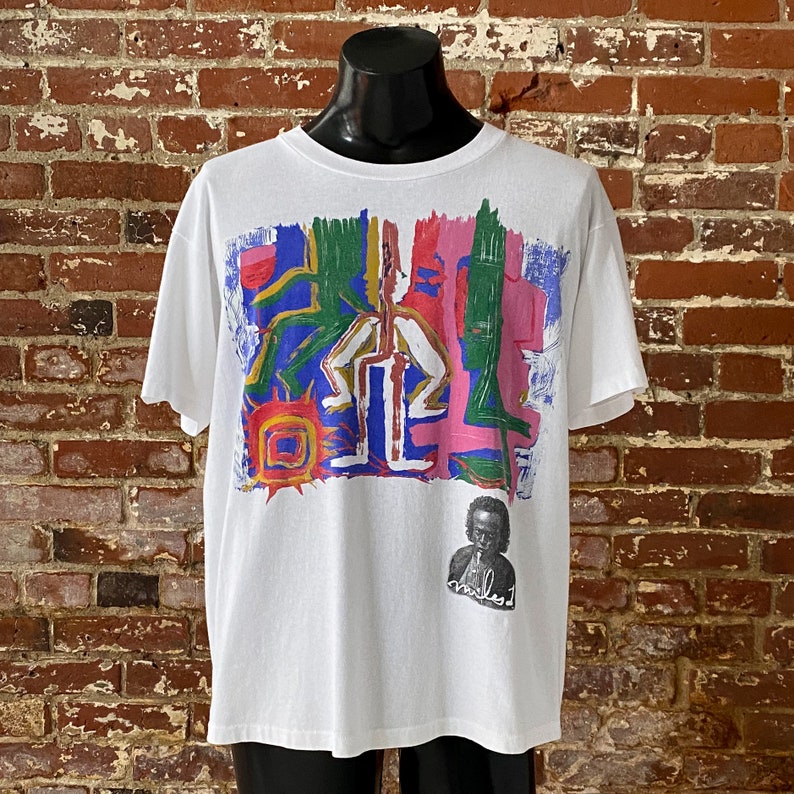 May include: White t-shirt with a colorful abstract design featuring a sun, figures, and lines. A black and white portrait of a man with the word "miles" written below is in the bottom right corner.
