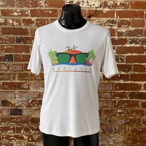 May include: White t-shirt with a graphic of Ray-Ban Wayfarer sunglasses with palm trees and a sunset. The text "Ray-Ban" and "Wayfarer" are printed on the shirt.