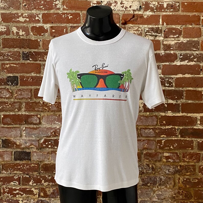 May include: White t-shirt with a graphic of Ray-Ban Wayfarer sunglasses with palm trees and a sunset. The text "Ray-Ban" and "Wayfarer" are printed on the shirt.