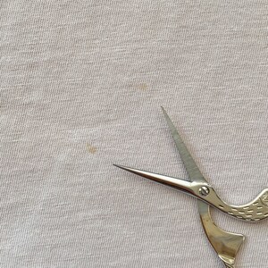 May include: A pair of silver-colored scissors with a bird-shaped handle. The scissors are on a white fabric background.