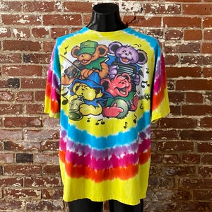 Op de afbeelding: Een tie-dye T-shirt met een gele achtergrond en een veelkleurig swirl-patroon. Het shirt heeft een afbeelding van vier cartoonberen die muziekinstrumenten bespelen. De beren dragen hoeden en hebben verschillende kleuren vacht. Het shirt heeft een ronde hals en korte mouwen.