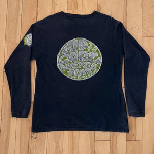 May include: A navy blue long-sleeve shirt with a large circular graphic on the back. The graphic features white text and illustrations on a green background. A smaller matching graphic is on the left sleeve.