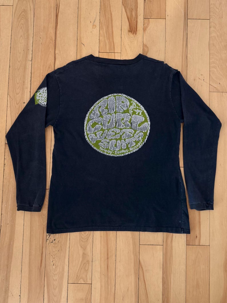 May include: A navy blue long-sleeve shirt with a large circular graphic on the back. The graphic features white text and illustrations on a green background. A smaller matching graphic is on the left sleeve.