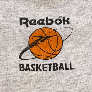 May include: A gray sweatshirt with the Reebok logo and the words "Basketball" printed on it. A basketball with an orange and black design is in the center of the sweatshirt.