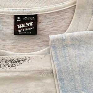 Puede incluir: Primer plano de una camiseta blanca con un dise&ntilde;o azul y negro descolorido. La camiseta tiene una etiqueta negra que dice "BEST FRUIT OF THE LOOM MADE IN U.S.A." y la talla es XL.
