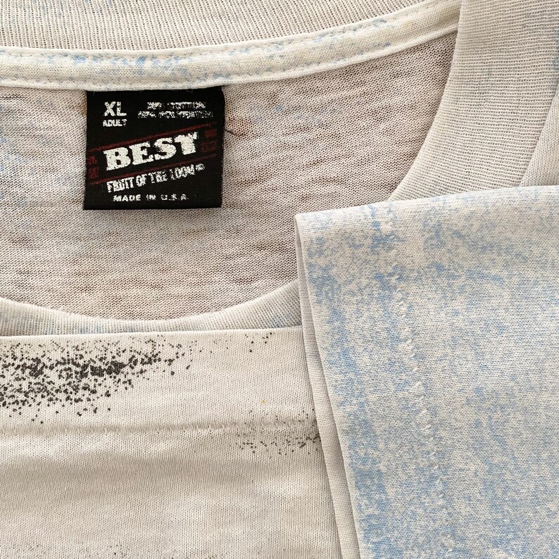 Puede incluir: Primer plano de una camiseta blanca con un dise&ntilde;o azul y negro descolorido. La camiseta tiene una etiqueta negra que dice "BEST FRUIT OF THE LOOM MADE IN U.S.A." y la talla es XL.