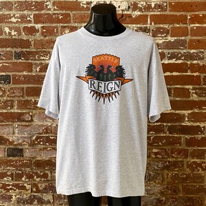 May include: Gray t-shirt with a graphic print of a crest featuring two black eagles with orange beaks and claws, a red castle, and the text "Seattle Reign".