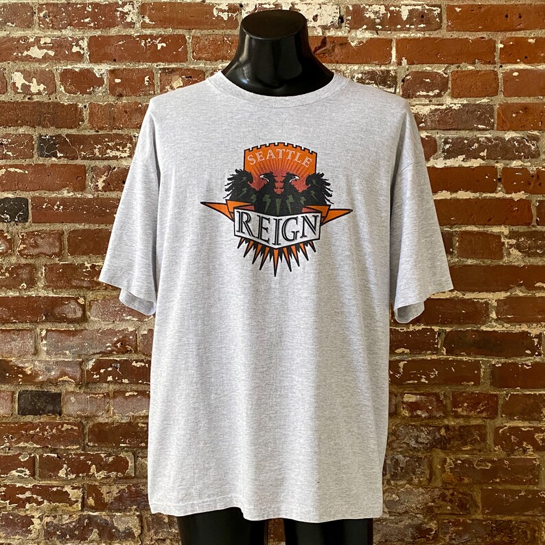 May include: Gray t-shirt with a graphic print of a crest featuring two black eagles with orange beaks and claws, a red castle, and the text "Seattle Reign".
