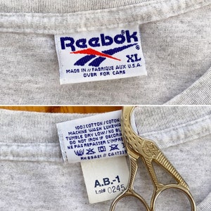 May include: Gray Reebok t-shirt with a white Reebok logo and size XL tag. The care instructions are printed on the tag. The tag also includes the text "Made in // Fabrique aux U.S.A.". A second tag is visible with the text "100% cotton/coton", "Machine wash luke warm", "Tumble dry low/no bleach", "Do not iron if decorated", "Ne pas repasser l'imprime", "RN 69421// CA17335", "A.B.-1", and "Lot# 0245".