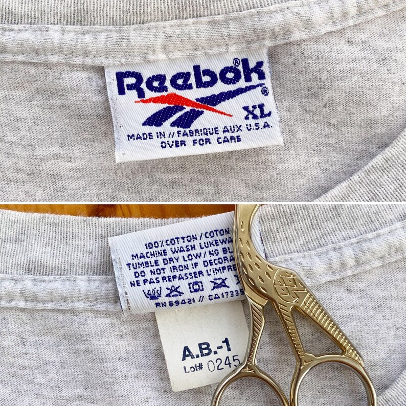 May include: Gray Reebok t-shirt with a white Reebok logo and size XL tag. The care instructions are printed on the tag. The tag also includes the text "Made in // Fabrique aux U.S.A.". A second tag is visible with the text "100% cotton/coton", "Machine wash luke warm", "Tumble dry low/no bleach", "Do not iron if decorated", "Ne pas repasser l'imprime", "RN 69421// CA17335", "A.B.-1", and "Lot# 0245".