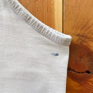 May include: A close-up of a light beige tank top with a small dark blue stain on the fabric.