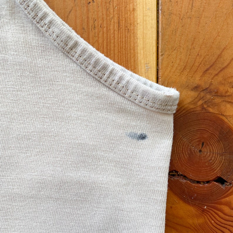 May include: A close-up of a light beige tank top with a small dark blue stain on the fabric.