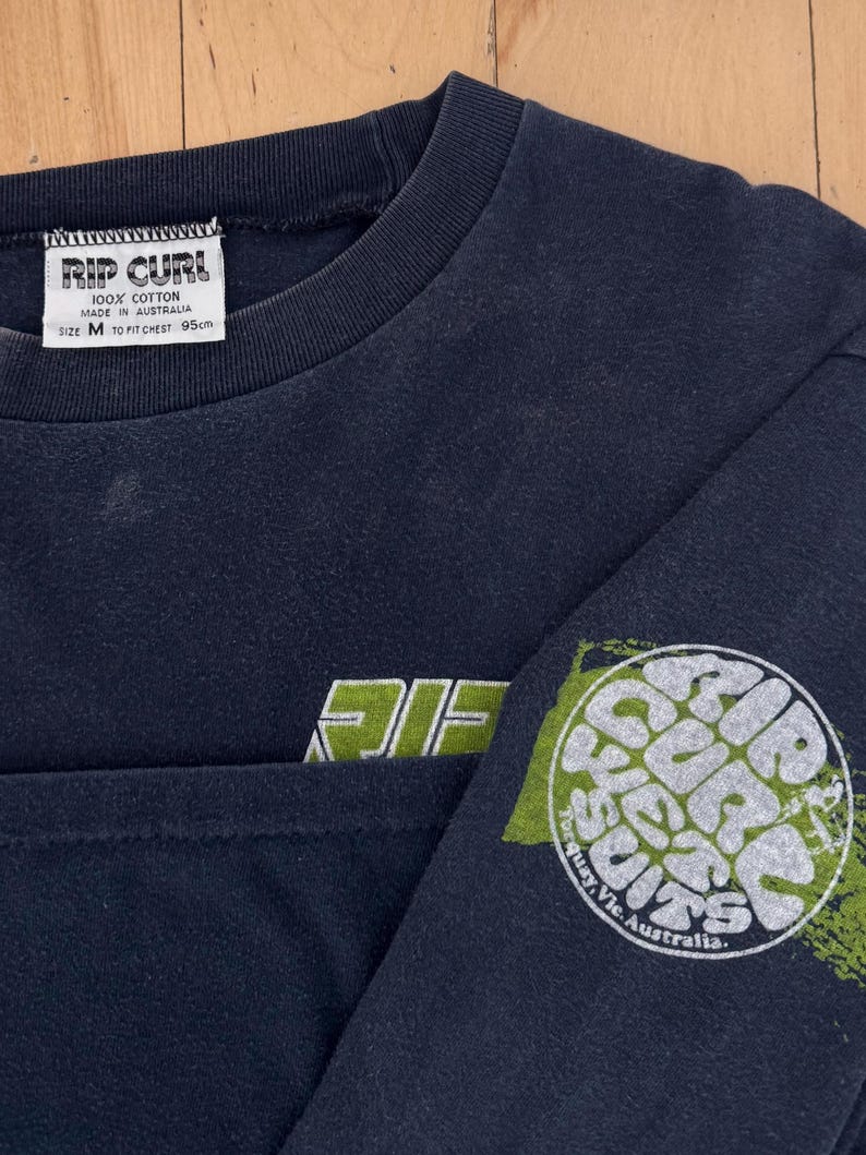 May include: Navy blue long-sleeve shirt with the text "RIP CURL" in white and green. The shirt has a crew neck and a graphic on the sleeve that says "RIP CURL" in a circular design. The shirt is size M, fits a 95cm chest, and is made of 100% cotton.
