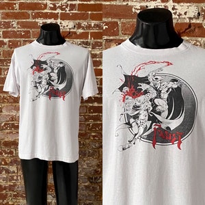 May include: White t-shirt with a black and white graphic print of a comic book character with a cape and a sword. The graphic is in a circle with the word "FAUST" printed below it.