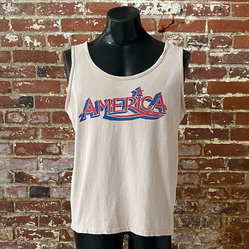 May include: A beige sleeveless tank top with a red, white, and blue graphic that says "AMERICA" with a star above the "A".