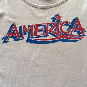 May include: A white tank top with a blue and red graphic that says "AMERICA" with two red stars above the word. The graphic has a blue wave design underneath the word.