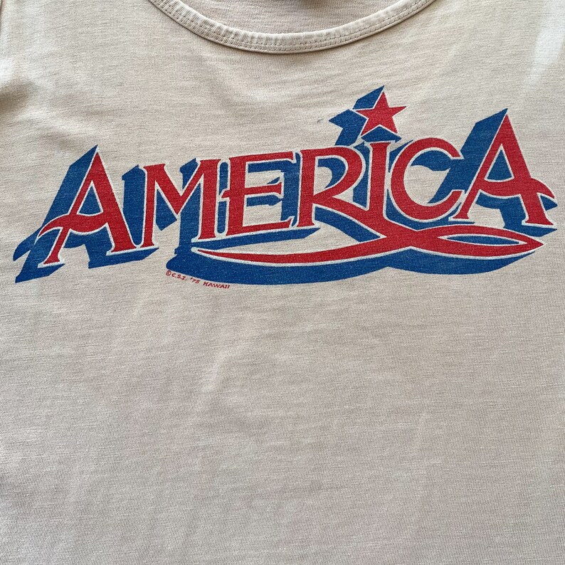 May include: A white tank top with a blue and red graphic that says "AMERICA" with two red stars above the word. The graphic has a blue wave design underneath the word.