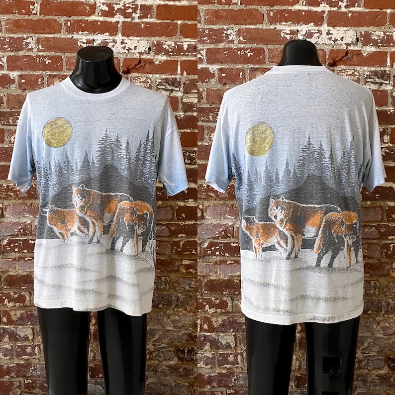 Puede incluir: Camiseta azul claro con un dise&ntilde;o de manada de lobos bajo la luna llena en un bosque. El dise&ntilde;o incluye lobos marrones y grises, una luna dorada y un paisaje nevado. La camiseta se muestra en un maniqu&iacute; negro.