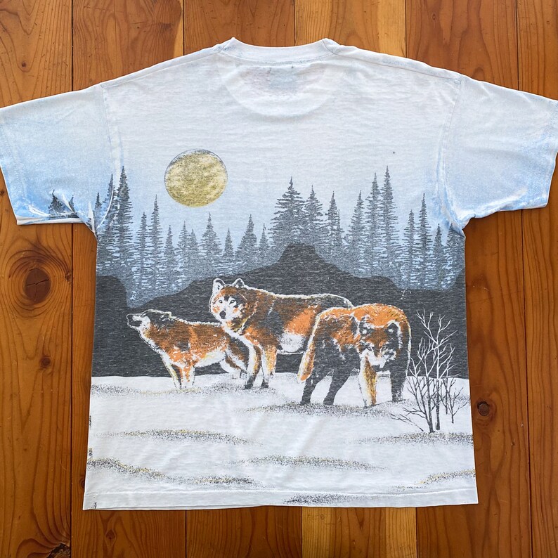 Puede incluir: Camiseta blanca con un dise&ntilde;o de manada de lobos en un bosque nevado bajo la luna llena. El dise&ntilde;o incluye tonos marrones, grises y azules, con ilustraciones detalladas de &aacute;rboles y lobos. La camiseta tiene cuello redondo.